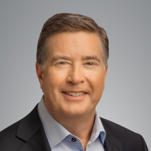 Studio headshot of a person(Michael Cole) wearing a dark blazer and light blue button-down shirt, positioned against a neutral gray background and facing forward.