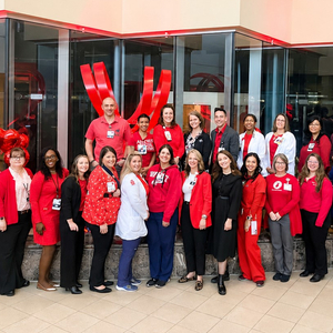 Dozens of physicians, nurses, and cardiovascular care team members from Sentara Heart Hospital gather for a group photo in the hospital atrium. Many are dressed in red and stand in front of a heart-themed display and artwork recognizing cardiovascular care.