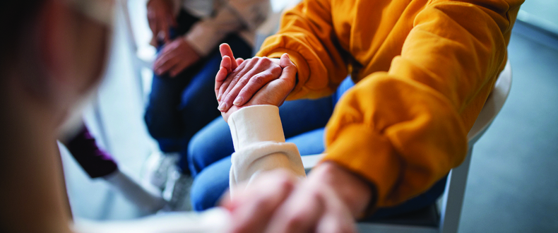 Close‑up of several adults seated in a circle holding hands during a group support or counseling session in an indoor setting.