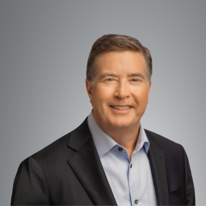 Studio headshot of a person wearing a dark blazer and light blue button-down shirt, positioned against a neutral gray background and facing forward.