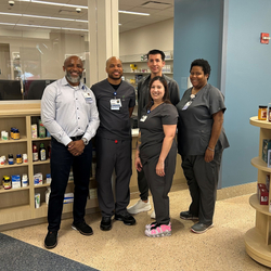 Five Sentara team members, including pharmacy staff, pose for a group photo inside the new Sentara To Home outpatient pharmacy at Sentara Northern Virginia Medical Center, surrounded by shelves of medications, health products and gifts.