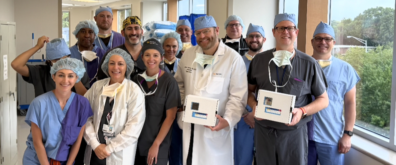 A group of 12 healthcare professionals poses together in a brightly lit hospital hallway lined with large windows. The team wears surgical scrubs in shades of blue, gray, and purple, along with surgical caps and some masks hanging around their necks. Two members near the front hold small white medical supply boxes, while another wears a white physician's coat.