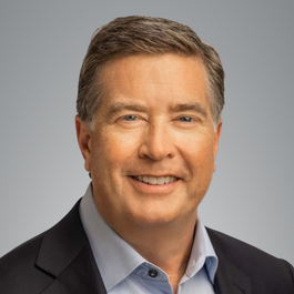 Studio headshot of a person wearing a dark blazer and light blue button-down shirt, positioned against a neutral gray background and facing forward.