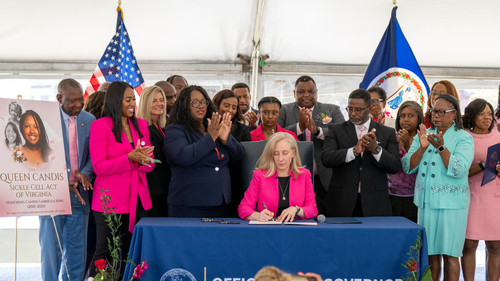 Virginia Governor Abigail Spanberger signs the Queen Candis Sickle Cell Act of Virginia during a ceremonial bill signing at the Sentara Community Care Center in Newport News, Thursday, June 11, 2026. Family members of Candis Gabriella King, elected officials, healthcare leaders, and community advocates stand behind her applauding, with Virginia and U.S. flags and a tribute display honoring Candis visible in the background.