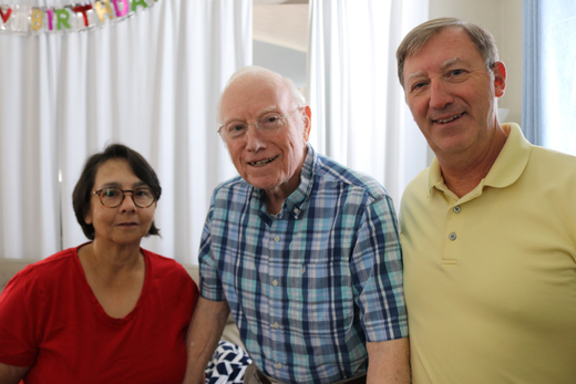 Three people pose closely together indoors beneath colorful “Happy Birthday” decorations. The person on the left wears a red shirt and glasses, the person in the center wears glasses and a blue plaid shirt, and the person on the right wears a pale yellow polo shirt. Bright windows and vertical blinds are visible in the background.
