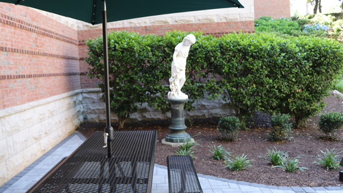 Outdoor hospital courtyard seating area with metal picnic table, green umbrella, landscaped shrubs, and a decorative statue against a brick and stone building wall.