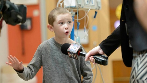 Child speaking into a news microphone inside a hospital room with medical equipment in the background.