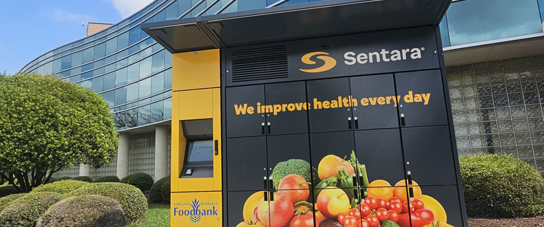Sentara community food access kiosk installed outside a modern glass building, featuring the Sentara logo, the message “We improve health every day,” and images of fresh fruits and vegetables, with Virginia Peninsula Foodbank branding visible.