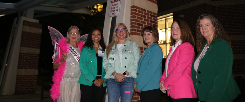 Six people stand together outside a brick building beneath a covered entrance illuminated with pink and teal lighting. One person wears decorative wings, a bright pink feather boa and a sash, while the other five wear jackets in shades of green, teal, light blue and bright pink.