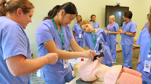 A Sentara team member demonstrates an airway management technique on a medical mannequin while middle school students wearing blue scrubs look on during Sentara's 2026 summer healthcare career camp at Sentara Williamsburg Regional Medical Center.