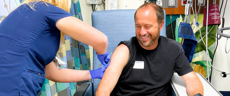A healthcare worker wearing gloves administers an injection to a seated patient in a hospital treatment room. Medical equipment and monitors are visible in the background.