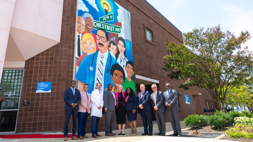Virginia Governor Abigail Spanberger stands with community and healthcare leaders outside the Sentara Community Care Center in Newport News beneath a colorful mural on the building's Chestnut Avenue facade. Joining her are Candis Gabriella King's mother and father, Newport News Mayor Phillip Jones, Sentara President and CEO Dennis Matheis, Sentara Chief Administrative Officer Aubrey Layne, Sentara Chief Clinical Officer Dr. Michael Hooper, Sentara Vice President of Government Relations Andy Stephenson, and other attendees following the ceremonial signing of the Queen Candis Sickle Cell Act, Thursday, June 11, 2026.