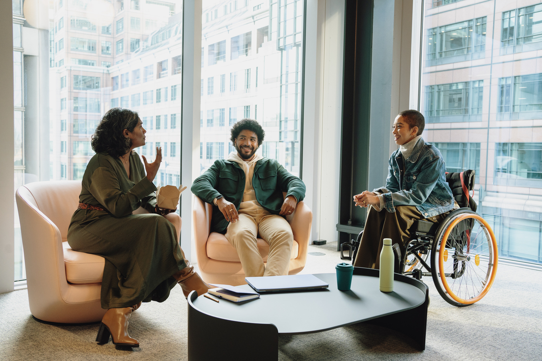 Three colleagues sit in a modern office lounge, including one person in a wheelchair, talking around a low coffee table with notebooks, a laptop, and drinks. Large windows behind them show tall city buildings..