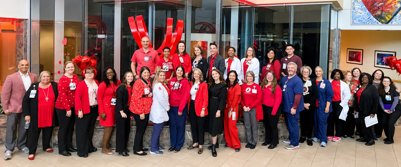 Dozens of physicians, nurses, and cardiovascular care team members from Sentara Heart Hospital gather for a group photo in the hospital atrium. Many are dressed in red and stand in front of a heart-themed display and artwork recognizing cardiovascular care.
