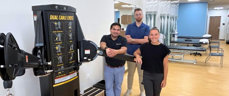 Three members of the Sentara Therapy Center team in Manassas, Virginia pose beside rehabilitation equipment inside the new facility on Liberia Avenue. Treatment tables and other physical therapy equipment are visible in the background.