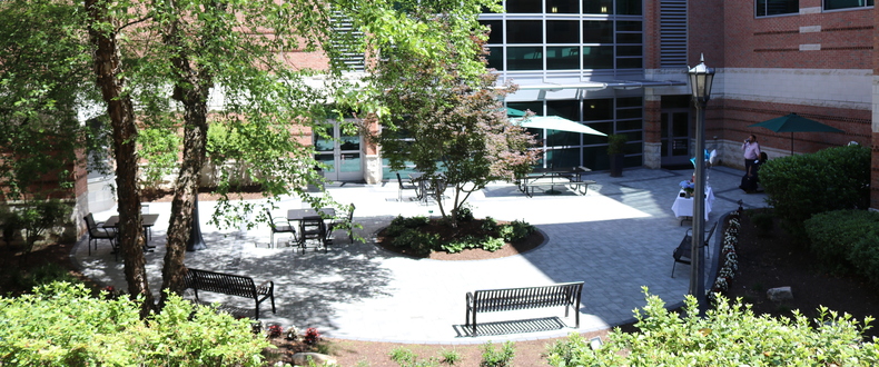 Landscaped courtyard with trees, benches, and outdoor seating outside a modern brick and glass hospital building, featuring a patio with tables, umbrellas, and walking paths.