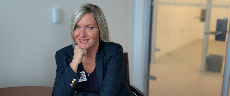 Professional portrait of a healthcare employee seated at a conference table in an office setting, wearing a blue plaid blazer and employee badge, with a glass doorway and hallway visible in the background.