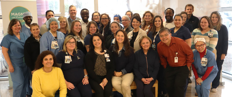 Members of Sentara Northern Virginia Medical Center’s MRSA prevention team gather for a group photo inside the hospital. Healthcare professionals and support staff wearing scrubs, business attire, and identification badges stand and sit together in a lobby area near large windows. A Magnet with Distinction sign is visible in the background.