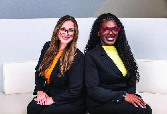 Two people in business attire seated side by side on a sofa in an office setting, facing the camera.