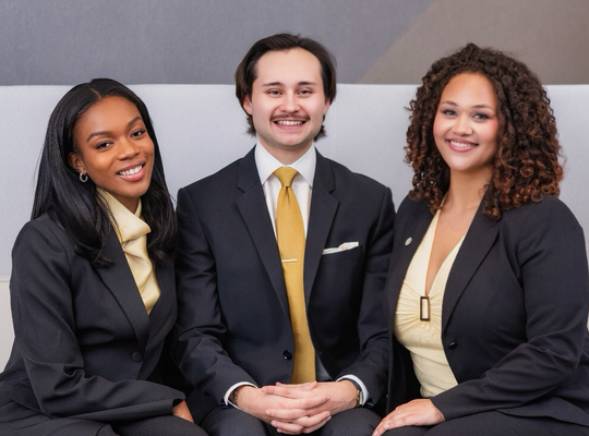 Three people in business attire seated together on a sofa in an office setting, facing the camera.