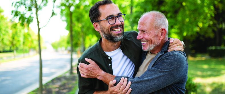 Two adults stand close together on a tree‑lined sidewalk, with one adult holding the other around the shoulders in an outdoor residential setting.