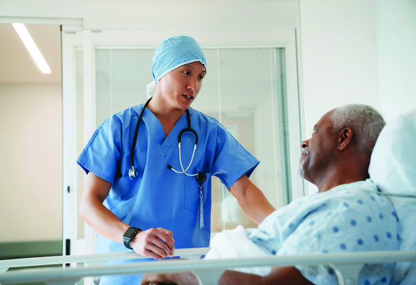 Healthcare professional standing beside a hospital bed and speaking with a patient in a clinical care setting.
