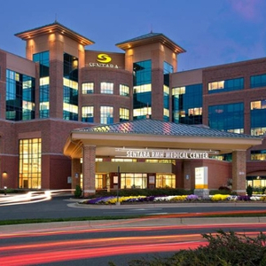 Exterior view of Sentara RMH Medical Center at dusk, featuring the illuminated hospital entrance, multi-story brick and glass facade, landscaped flower beds, and light trails from passing vehicles in Harrisonburg, Virginia.