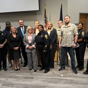 Group photo of several public safety officers and community representatives standing together indoors in front of U.S. and state flags.