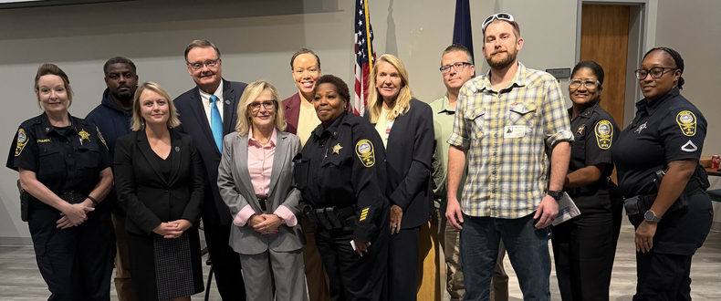 Group photo of several public safety officers and community representatives standing together indoors in front of U.S. and state flags.