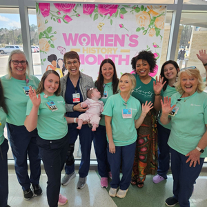 Nine labor and delivery healthcare workers pose in front of a Women's History Month banner at Sentara Albemarle Medical Center. The middle worker is holding an infant.