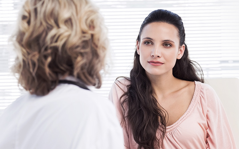 Person seated facing a medical professional in a clinical setting during a consultation, with both individuals visible from the shoulders up.
