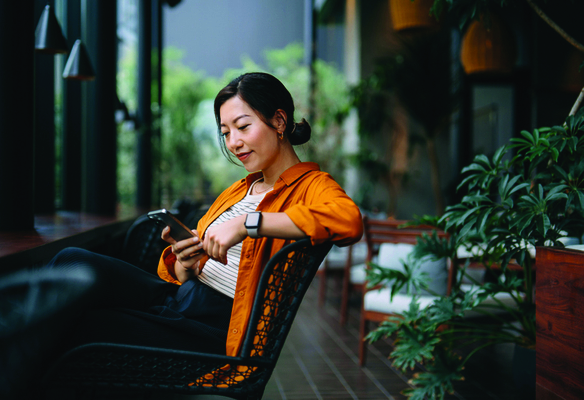 Person using a smartphone while seated on a patio surrounded by plants in an outdoor lounge setting.