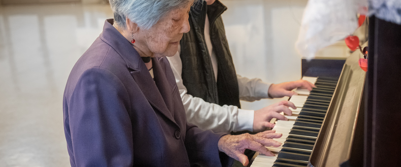 Older adult playing a piano while another adult stands nearby in an indoor room.
