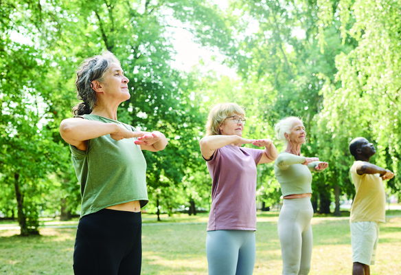 Four people practice tai chi outdoors in a park-like setting, standing in a line with arms raised at chest level in a slow, controlled posture. The group wears casual exercise clothing, and leafy green trees fill the background, illustrating a gentle, low-impact tai chi session focused on balance, posture, and mindful movement.