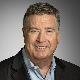 Professional headshot of a person wearing a dark blazer and light blue collared shirt, facing the camera against a gray studio background.