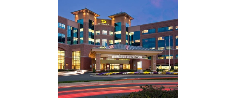 Exterior view of Sentara RMH Medical Center at dusk, featuring the illuminated hospital entrance, multi-story brick and glass facade, landscaped flower beds, and light trails from passing vehicles in Harrisonburg, Virginia.