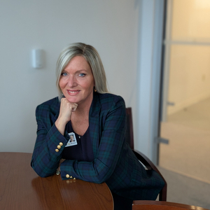 Professional portrait of a healthcare employee seated at a conference table in an office setting, wearing a blue plaid blazer and employee badge, with a glass doorway and hallway visible in the background.