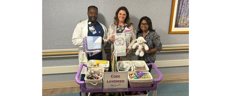 Three healthcare team members stand behind a purple Code Lavender support cart in a hospital hallway. The cart contains comfort items, books, sensory tools, snacks, a stuffed lamb, and a folded quilt used to support patients, families, and caregivers during stressful situations. A sign on the cart reads “Code Lavender.”