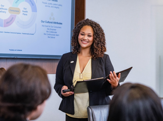 Presenter standing in a meeting room in front of a presentation screen, addressing attendees.