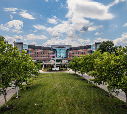 Exterior view of a Sentara hospital building with a landscaped lawn, trees, walkways, and an American flag in front of the main entrance.