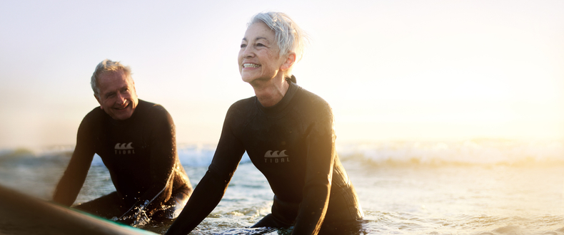 Two adults in wetsuits sit on surfboards in shallow ocean water during sunset, with waves and warm sunlight visible in the background.