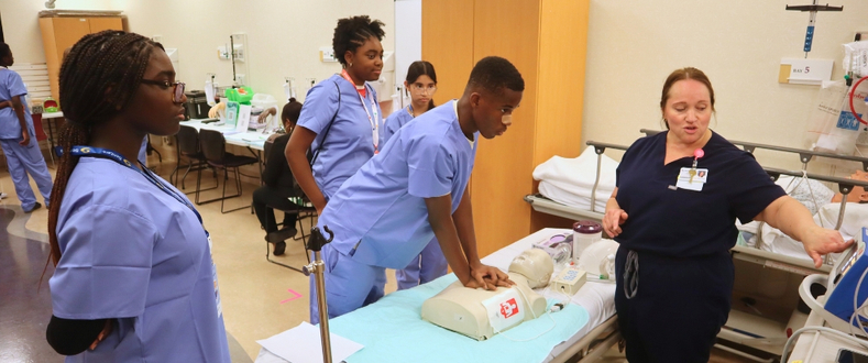 A Sentara team member guides middle school students wearing blue scrubs through a CPR demonstration on a mannequin during Sentara's 2026 summer healthcare career camp at Sentara Northern Virginia Medical Center.