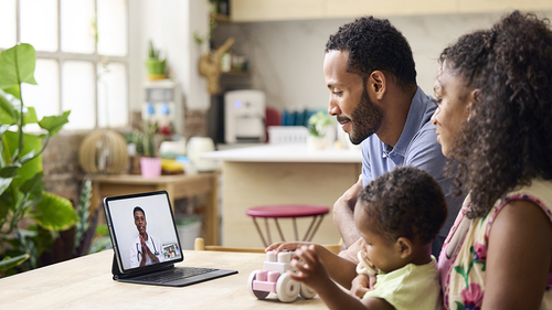 Family sitting at a dining table using a tablet for a video call with a healthcare professional, while a young child plays with a toy in a bright kitchen setting.