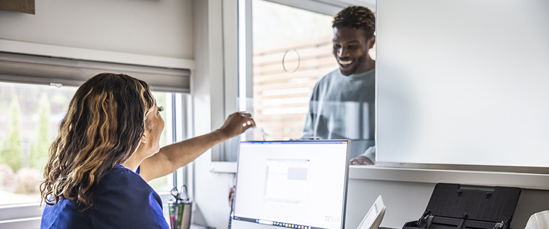 Person at a front desk using a computer hands a card through a service window to a visitor, illustrating patient check‑in at a healthcare reception area.