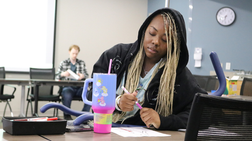 A participant in a support group activity sits at a conference table drawing on a body outline worksheet while other attendees work in the background.
