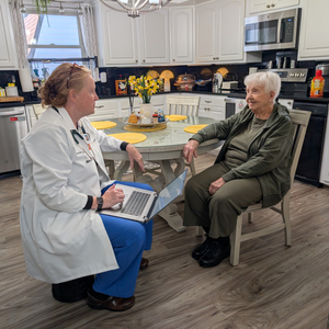 Healthcare professional seated with an older adult at a kitchen table, using a laptop during an in‑home visit.