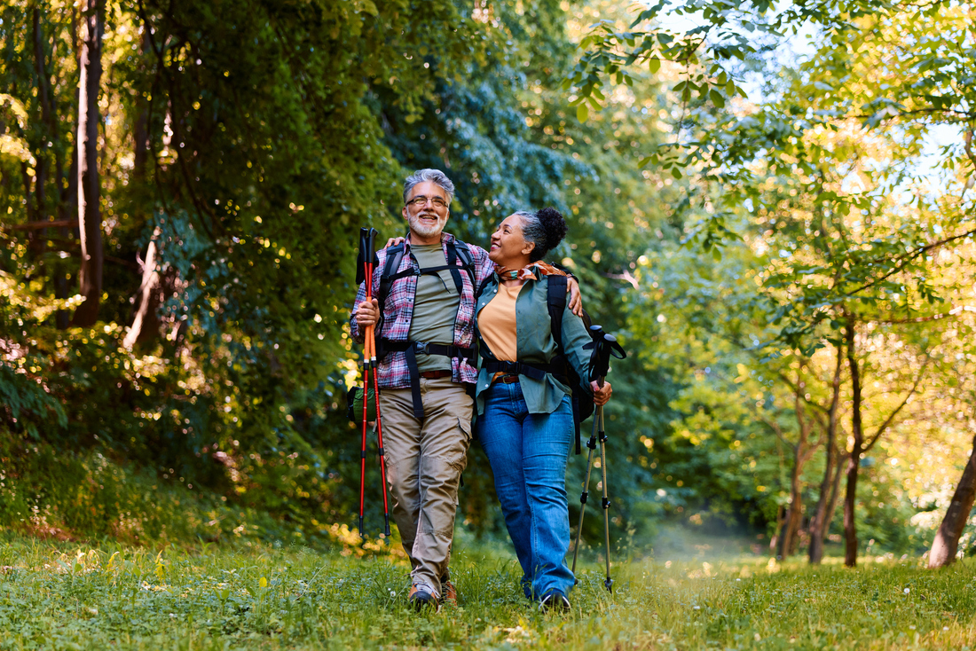 Two adults wearing backpacks and using trekking poles walk together along a grassy path through a wooded area. Tall trees and green foliage surround the trail, with sunlight filtering through the leaves overhead.