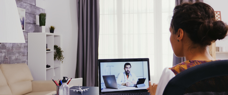 Woman participating in a telehealth video consultation with a doctor on a laptop at a home office desk.