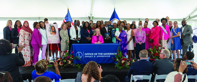 Virginia Governor Abigail D. Spanberger sits at a signing table draped with an Office of the Governor banner, surrounded by dozens of elected officials, community advocates, and health care leaders gathered under a tent for the signing of the Queen Candis Sickle Cell Act of Virginia. A commemorative poster honoring Queen Candis is held up on the left side of the group. The American and Virginia state flags are visible in the background, and floral arrangements line the front of the stage.