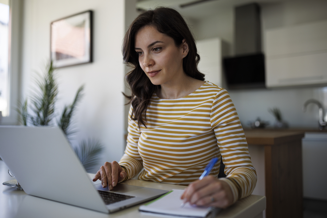 Person sitting at a desk in a modern home kitchen, typing on a laptop and writing in a notebook, wearing a yellow‑and‑white striped shirt.
